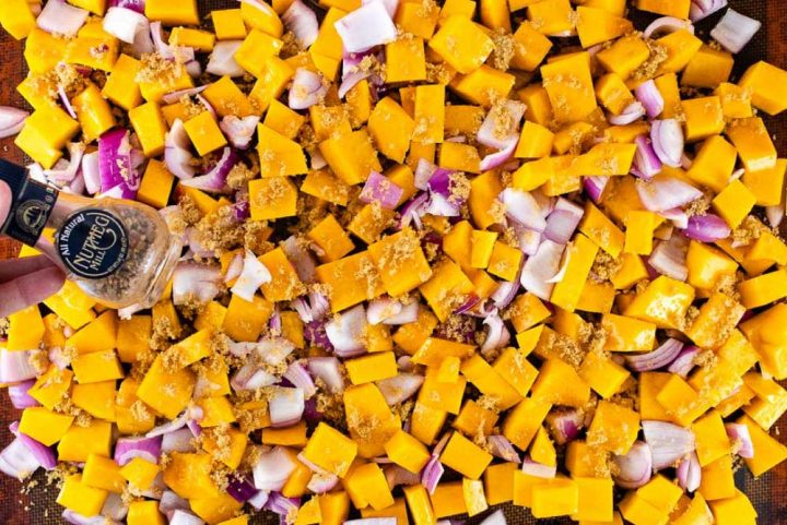 Sheet pan with cubed butternut squash and diced onion spread out and sprinkled with brown sugar; a jar with pieces of nutmeg is displayed above the pan