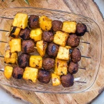 Glass dish with grilled vegetarian meatball and pineapple kabobs on a cutting board with whole pineapple to the side