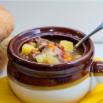 Brown and white soup bowl with vegetable beef soup and a spoon, next to a loaf of bread.