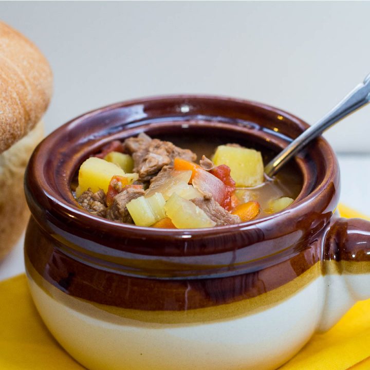 Brown and white soup bowl with vegetable beef soup and a spoon, next to a loaf of bread.