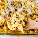 Side view of penne pasta bake topped with melted cheese in a glass pan with a spoonful scooped out and held toward the camera