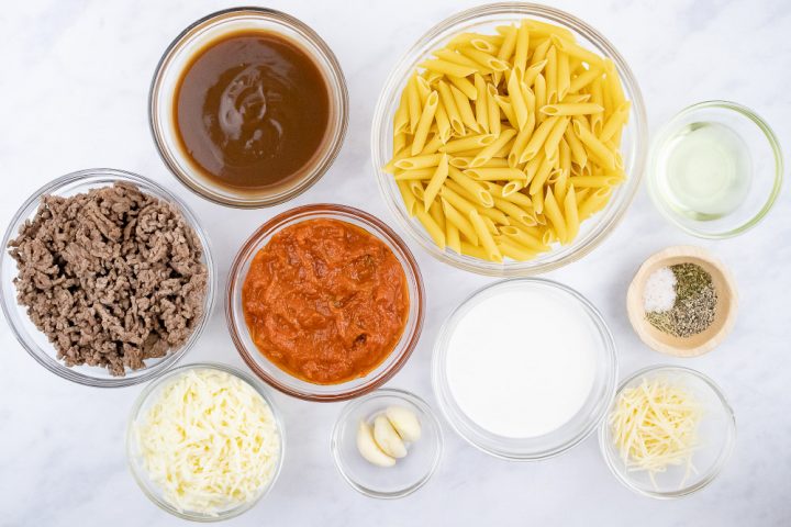 Overhead view of ingredients for Penne Pasta Bake: penne pasta, cooked ground beef, spaghetti sauce, brown gravy, garlic, half and half, mozzarella cheese, parmesan cheese, herbs, salt & pepper