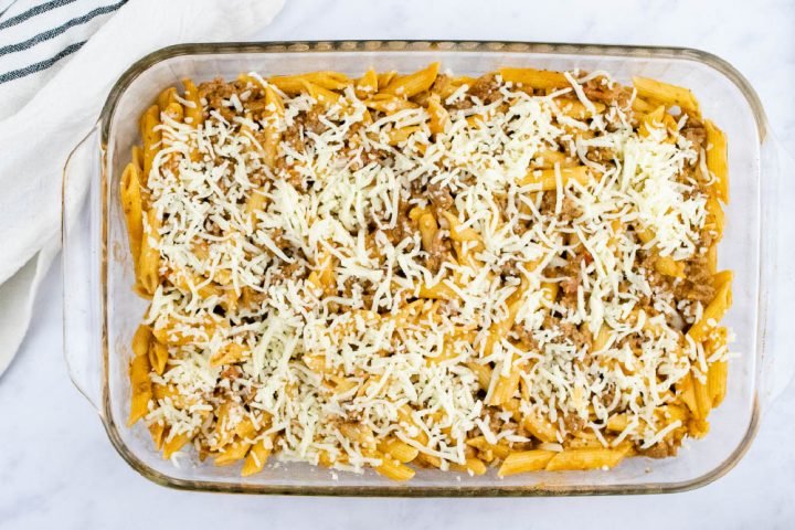 Overhead view of unbaked penne pasta bake topped with shredded cheese in a glass pan