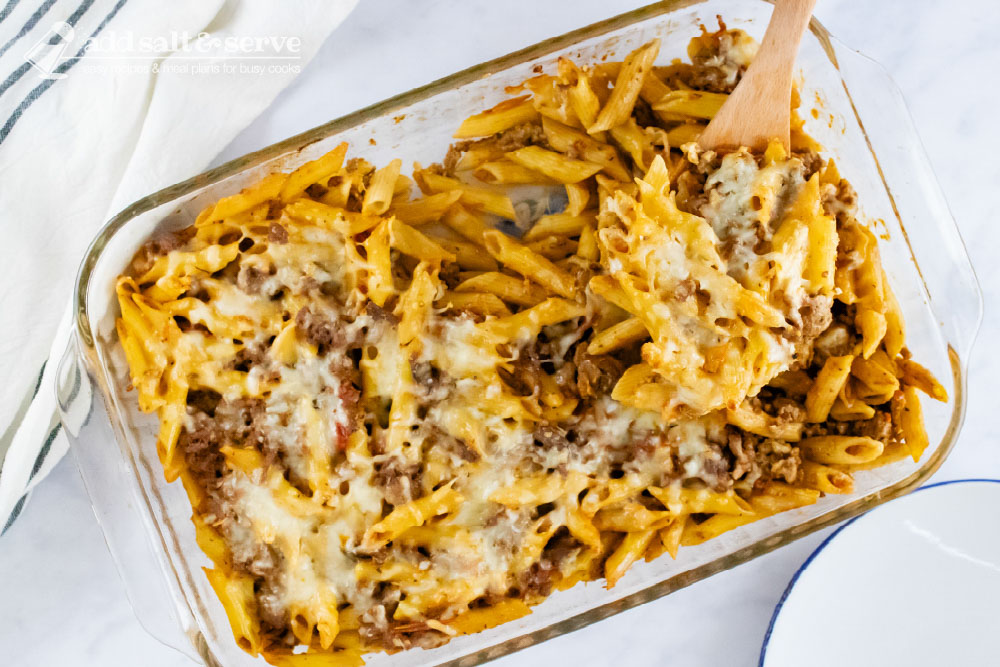 Overhead view of penne pasta bake topped with melted cheese in a glass pan with a spoonful scooped out and held toward the camera.