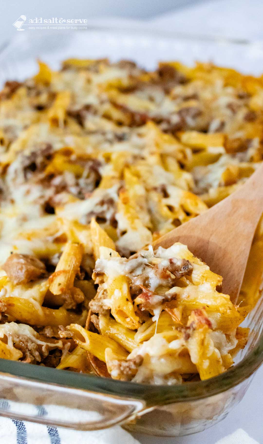Overhead view of penne pasta bake topped with melted cheese in a glass pan with a spoonful scooped out and held toward the camera.