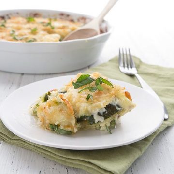 A serving of Cheezy Zucchini Casserole on a plate with a casserole dish full of the remaining recipe in the background