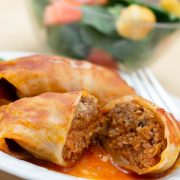 Closeup of cabbage rolls in a tomato sauce on a white plate with a fork. Bowl of salad in the background.