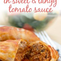 Closeup of cabbage rolls in a tomato sauce on a white plate with a fork. Bowl of salad in the background. Text is Cabbage Rolls in Sweet & Tangy Tomato Sauce - Add Salt & Serve logo