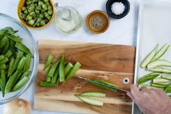 A bowl of okra and okra on a cutting board, with bowls of oil, salt, and pepper