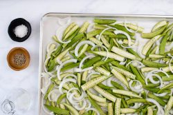 Chopped okra and onion on a baking sheet, nearby are bowls of oil, salt, and pepper