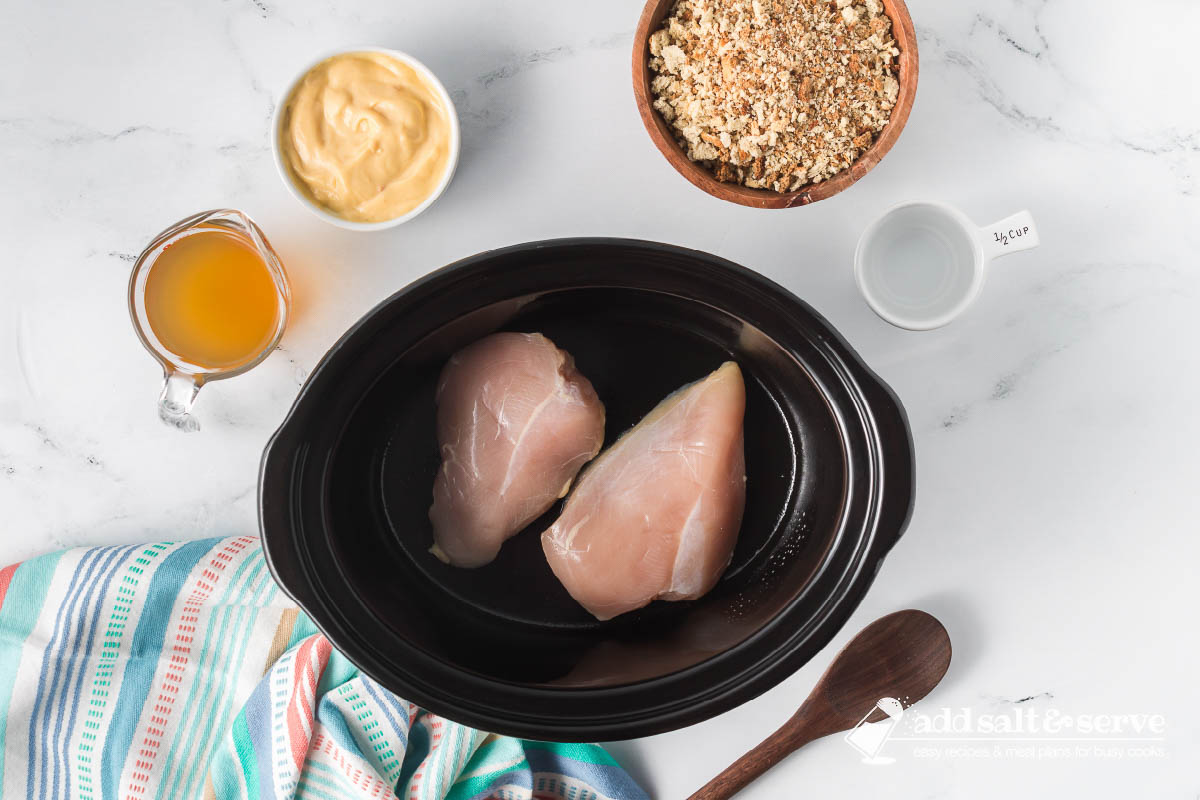 Two uncooked boneless chicken breasts in a crockpot surrounded by a bowl of dressing mix, a bowl of cream of chicken soup, a measuring cup of chicken broth, and a measuring cup of water.
