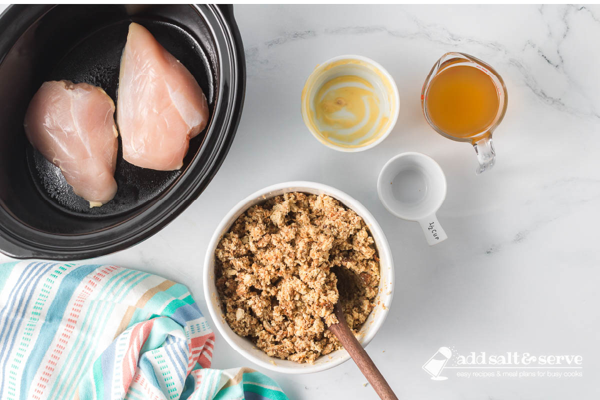 Two uncooked boneless chicken breasts in a crockpot surrounded by a bowl of dressing mix that has been combined with cream of chicken soup, a measuring cup of chicken broth, and a measuring cup of water.