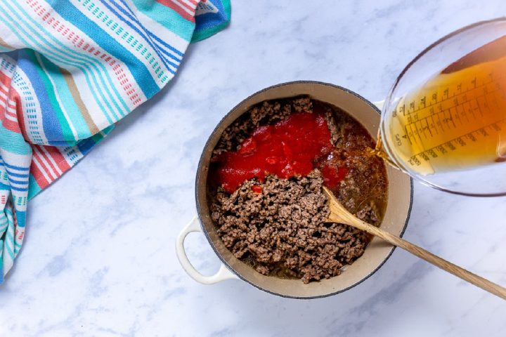 adding tomato sauce and beef broth to cooked ground beef in a dutch oven
