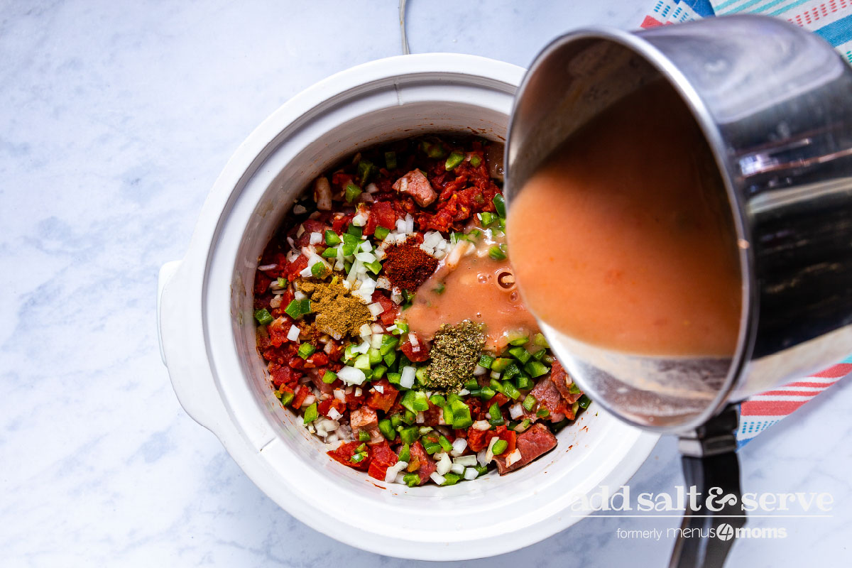 Pouring thickened sauce for Carne Guisada over meat, vegetables, and spices in crock pot.
