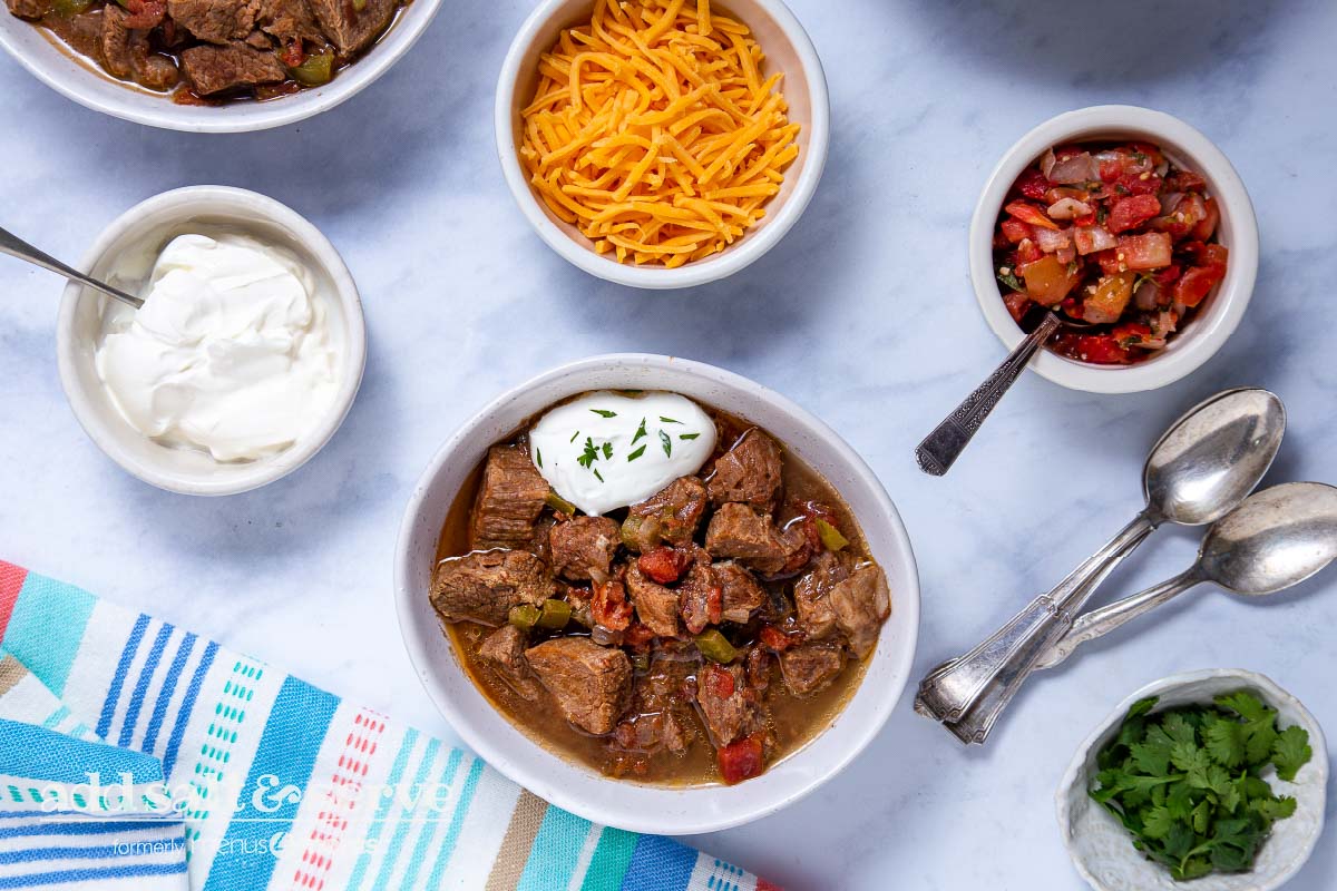Overhead view of Carne Guisada made in crock pot with garnishes in small bowls.