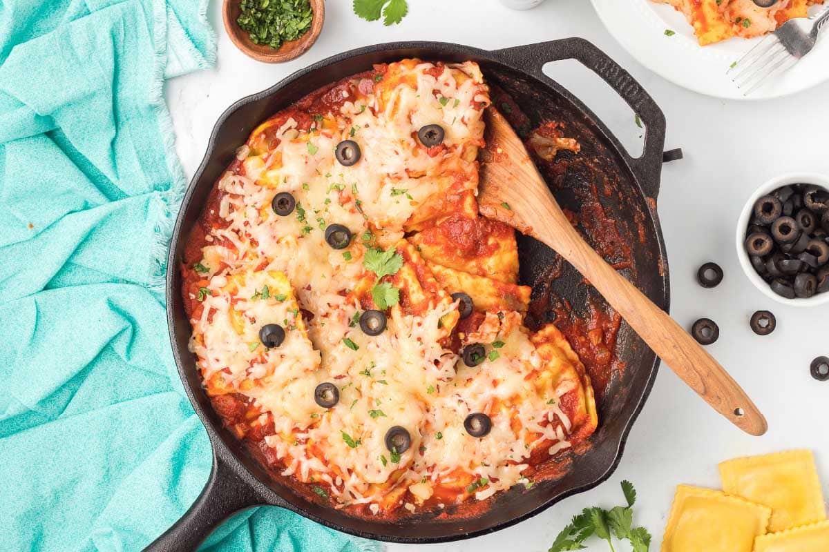 Cast iron skillet with cooked ravioli mixed with salsa, herbs, and tomato sauce and topped with cheese, black olives, and cilantro.