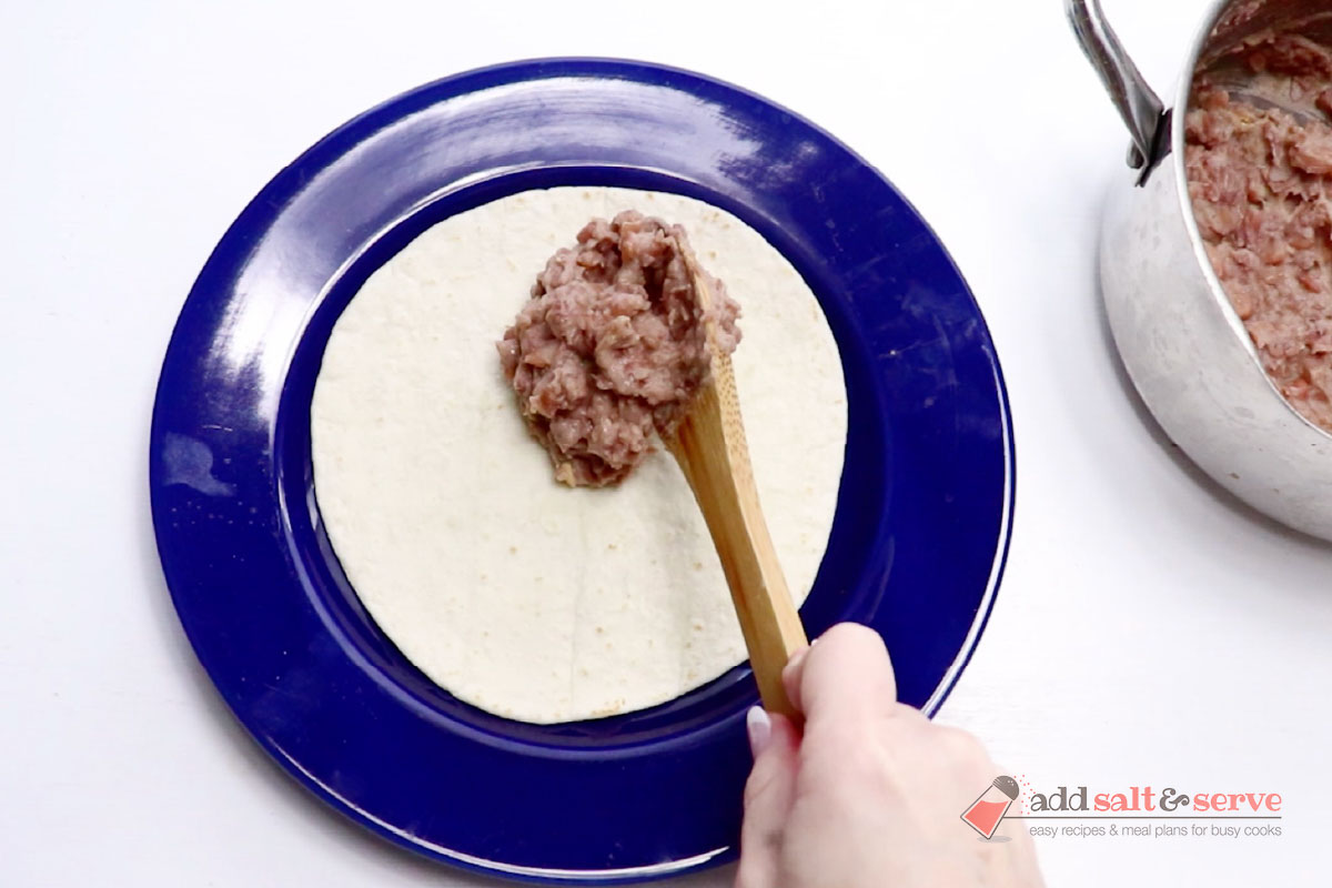 A tortilla on a plate with a spoonful of bean burrito filling being placed on it.
