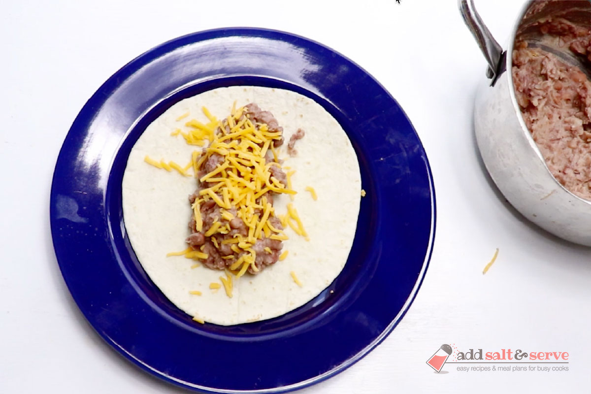 A tortilla on a plate with bean burrito filling and shredded cheddar cheese before folding into a burrito.