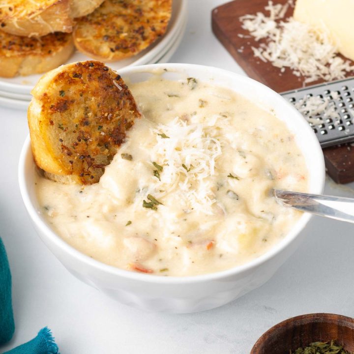 A bowl of Copycat Olive Garden Chicken Gnocchi Soup served with a piece of toasted garlic bread and a wedge of Parmesan cheese with a grater to the side.