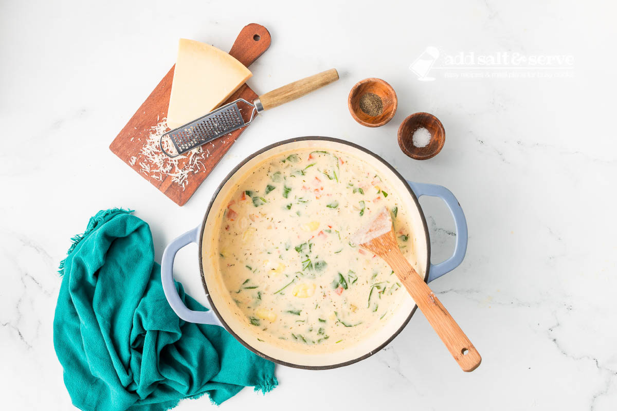 Copycat Olive Garden Chicken Gnocchi Soup in an enamel dutch oven with a wooden spoon in it and a wedge of Parmesan cheese, salt, and pepper beside the bowl.