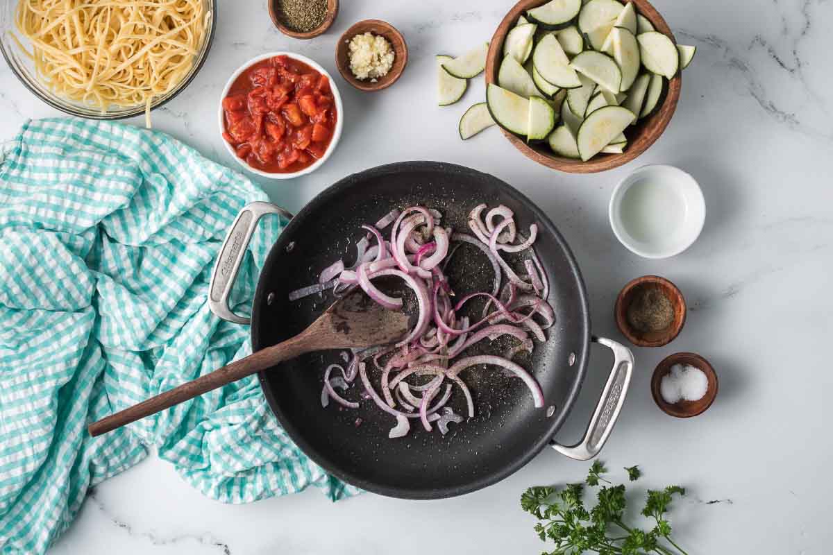 Sliced red onion sautéing in a skillet surrounded by dishes of sliced zucchini, cooked linguine, minced garlic, Italian seasoning, salt, pepper, and diced canned tomatoes.
