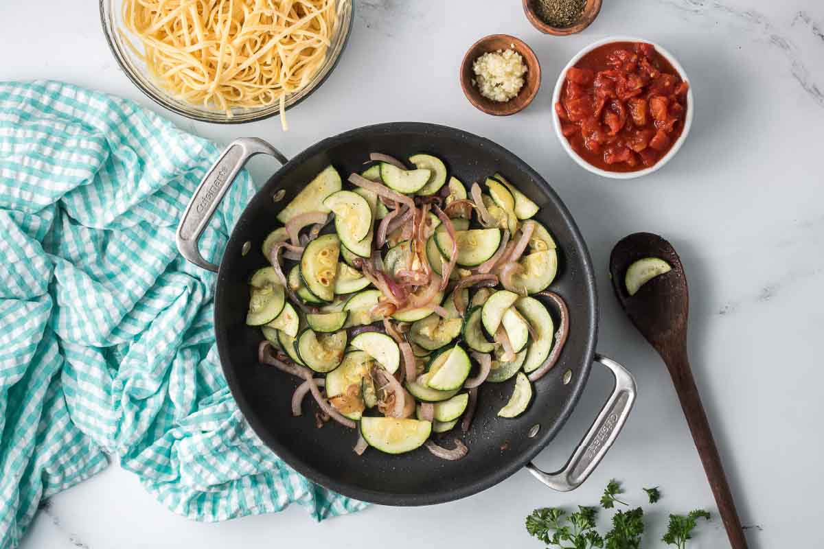 Sliced zucchini and red onion sautéing in a skillet surrounded by dishes of cooked linguine, minced garlic, Italian seasoning, and diced canned tomatoes.