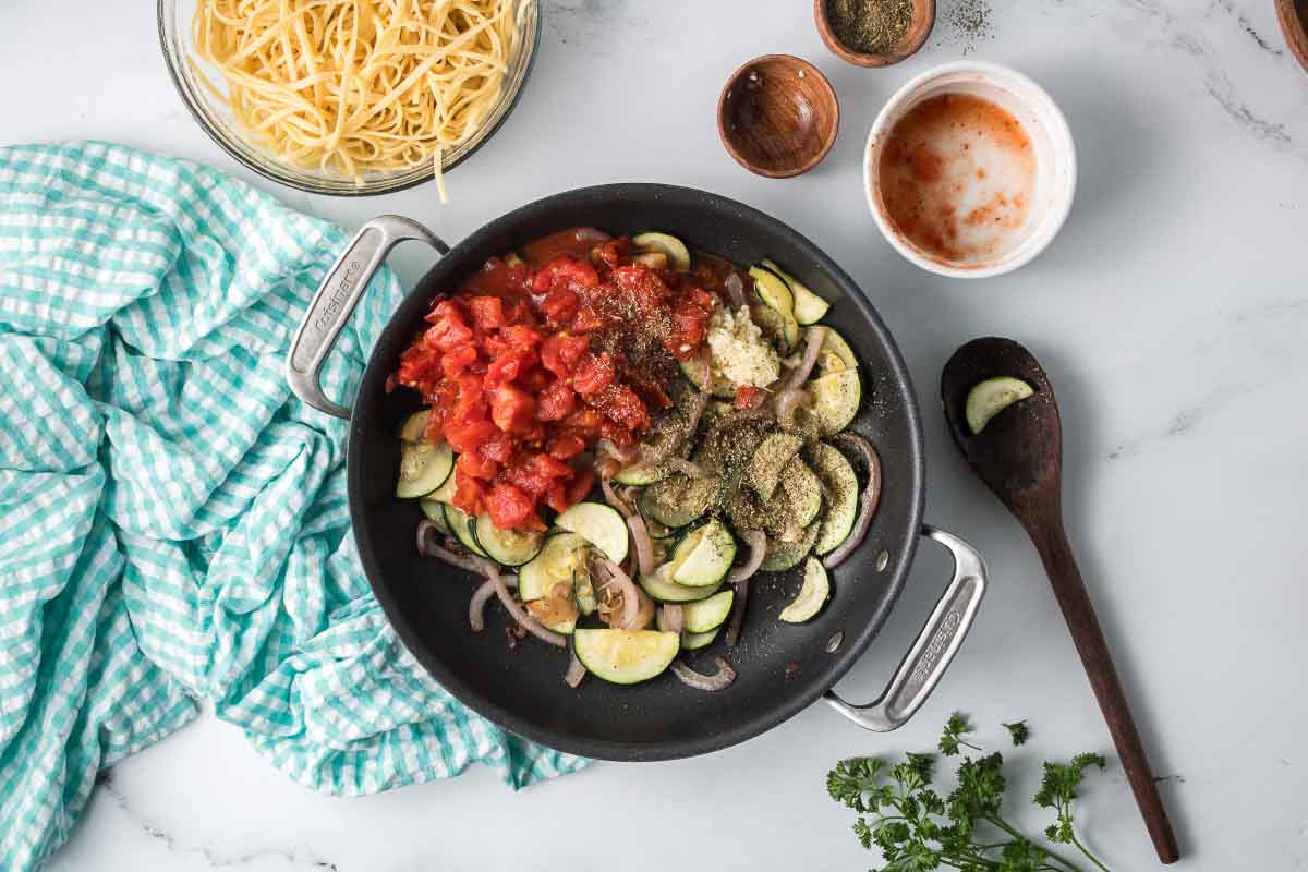 Sautéed sliced zucchini and red onion in a skillet with minced garlic, diced canned tomatoes, and Italian seasoning surrounded by dishes of cooked linguine and pepper.