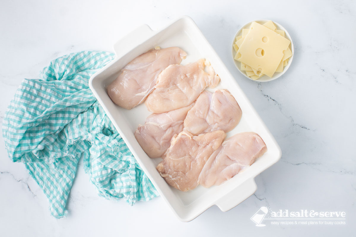 Casserole dish with chicken breasts and a plate with slices of Swiss cheese to the side.