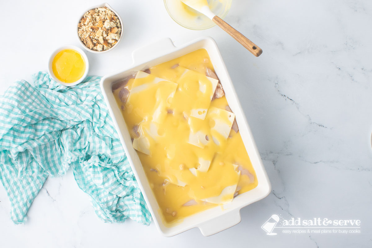 Casserole dish of chicken breasts and Swiss cheese with white wine sauce before adding stuffing and melted butter.
