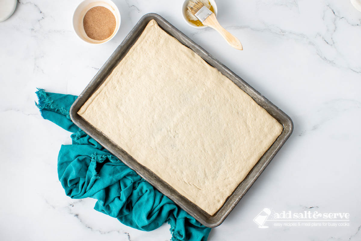 Pizza crust dough rolled out on a baking pan with melted butter and cinnamon sugar on the side.