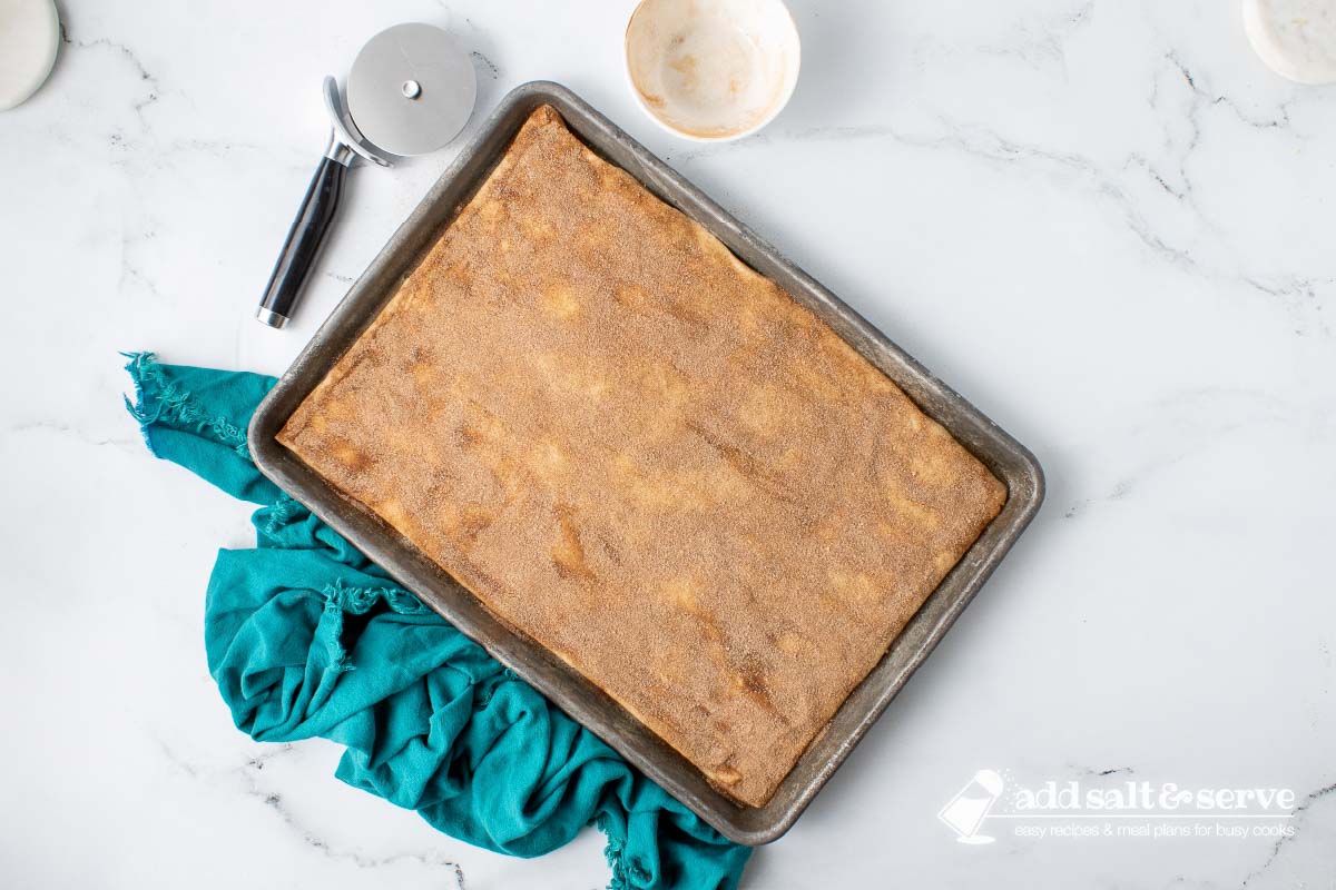 Pizza crust dough rolled out on a baking sheet and covered in melted butter and cinnamon sugar before baking.