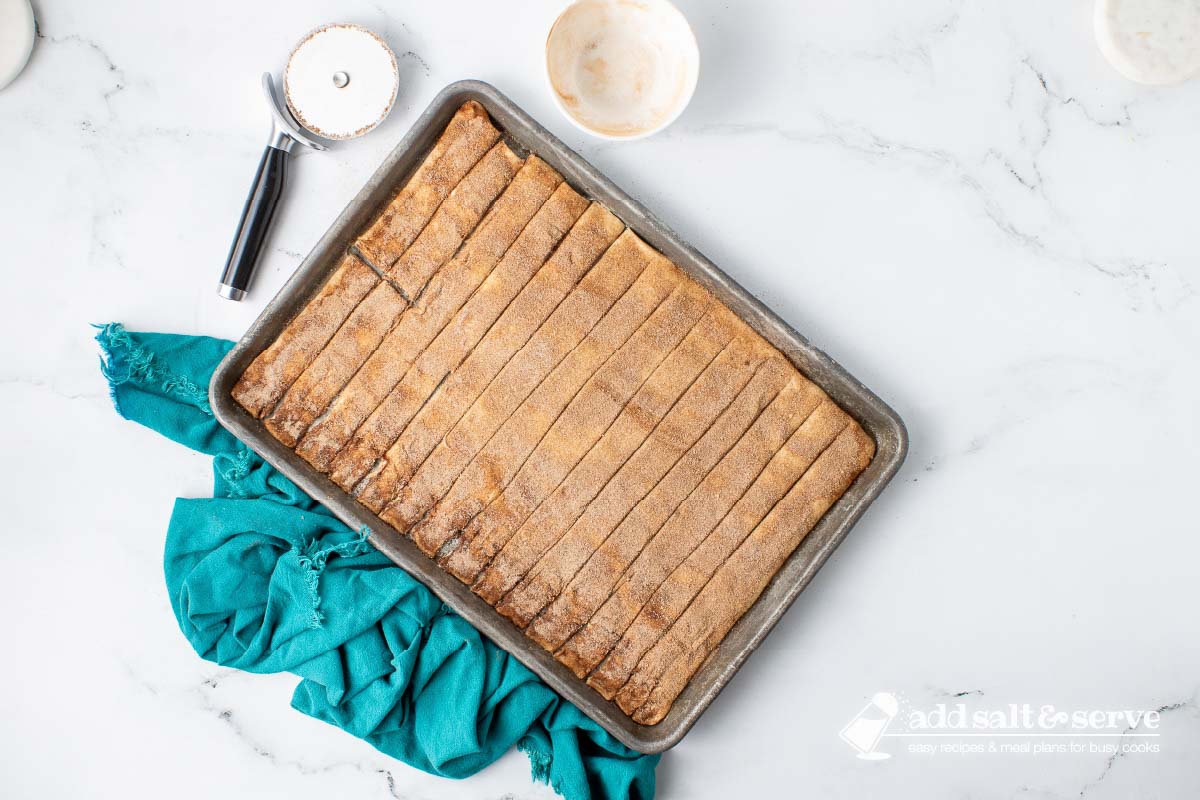 Cinnamon sticks topped with butter and cinnamon sugar on a baking sheet before baking.