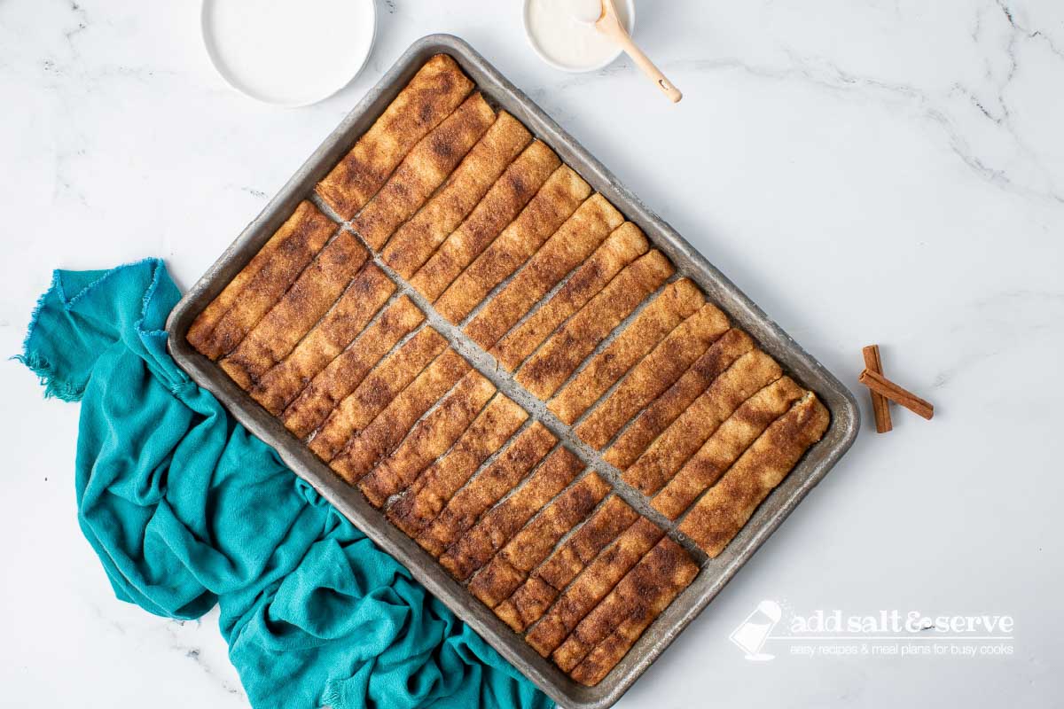 Baked cinnamon sticks encrusted with cinnamon sugar in butter on a baking sheet.