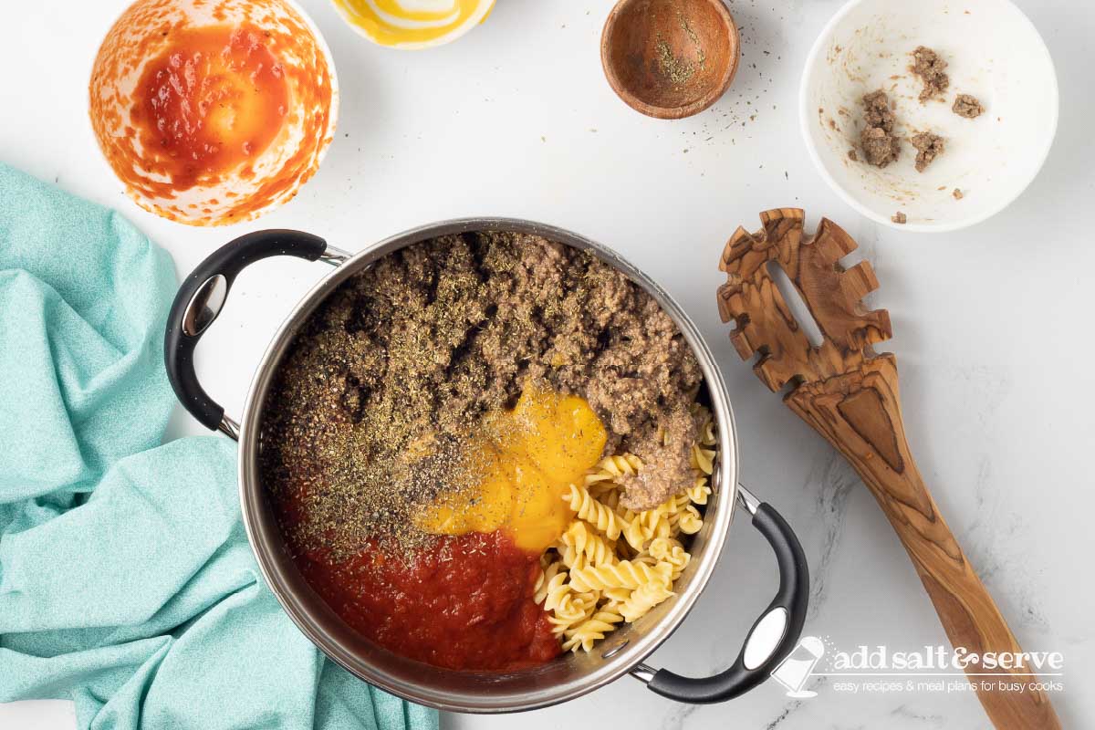 Large pot with cooked spiral pasta, cooked ground beef, marinara sauce, condensed cheddar cheese soup, Italian seasoning, and black pepper not yet mixed together.
