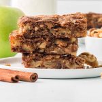 Three Apple Walnut Bars stacked on a white plate with a bowl of walnuts and a green apple in the background.