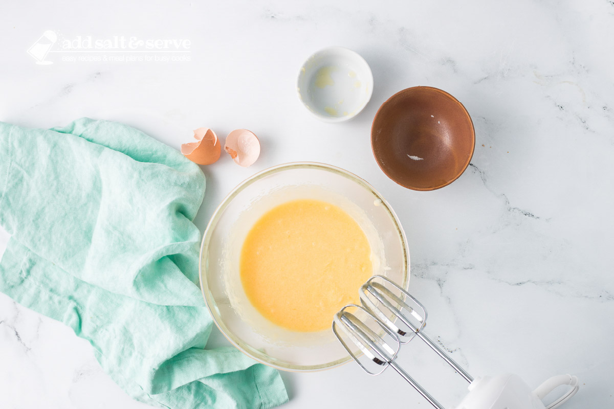 A clear glass bowl with a beaten mixture of melted butter, an egg, and sugar with the blender sitting to the side.