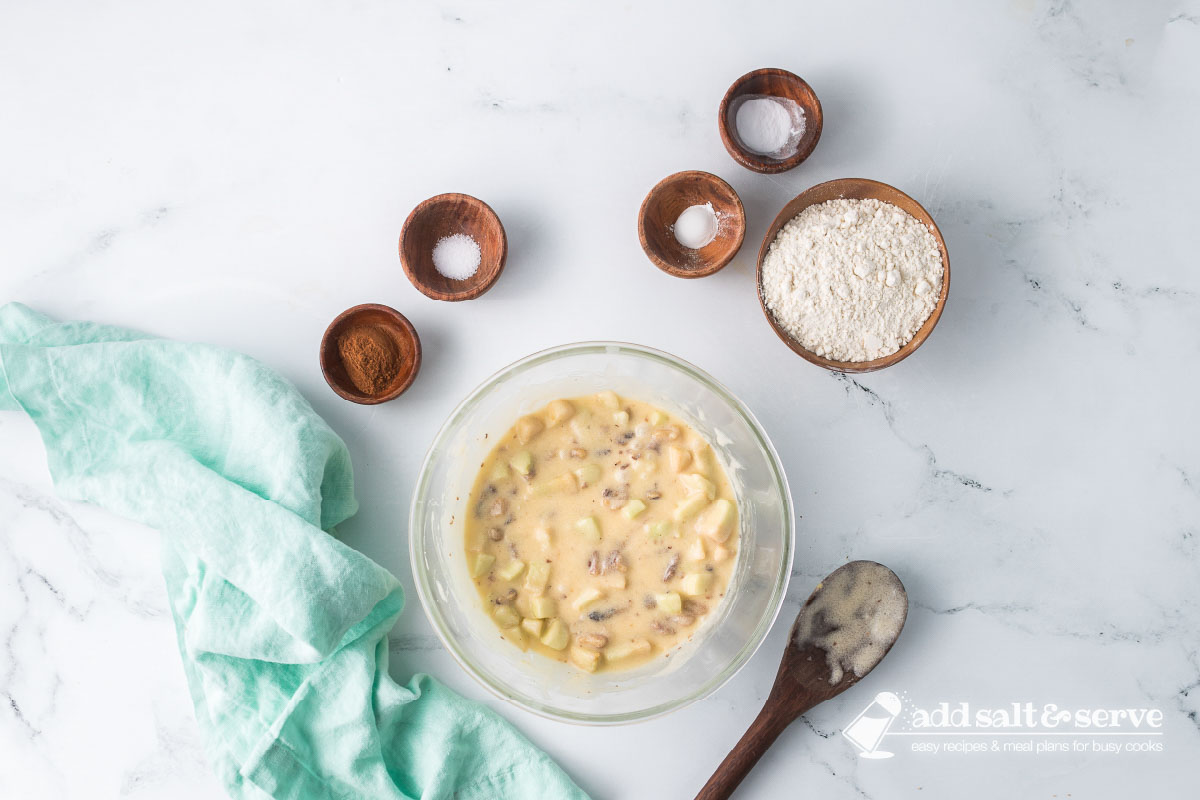 A clear glass bowl with beaten melted butter, an egg, and sugar with chopped apples and walnuts added and stirred in and flour, baking soda, baking powder, salt, and ground cinnamon to the side.