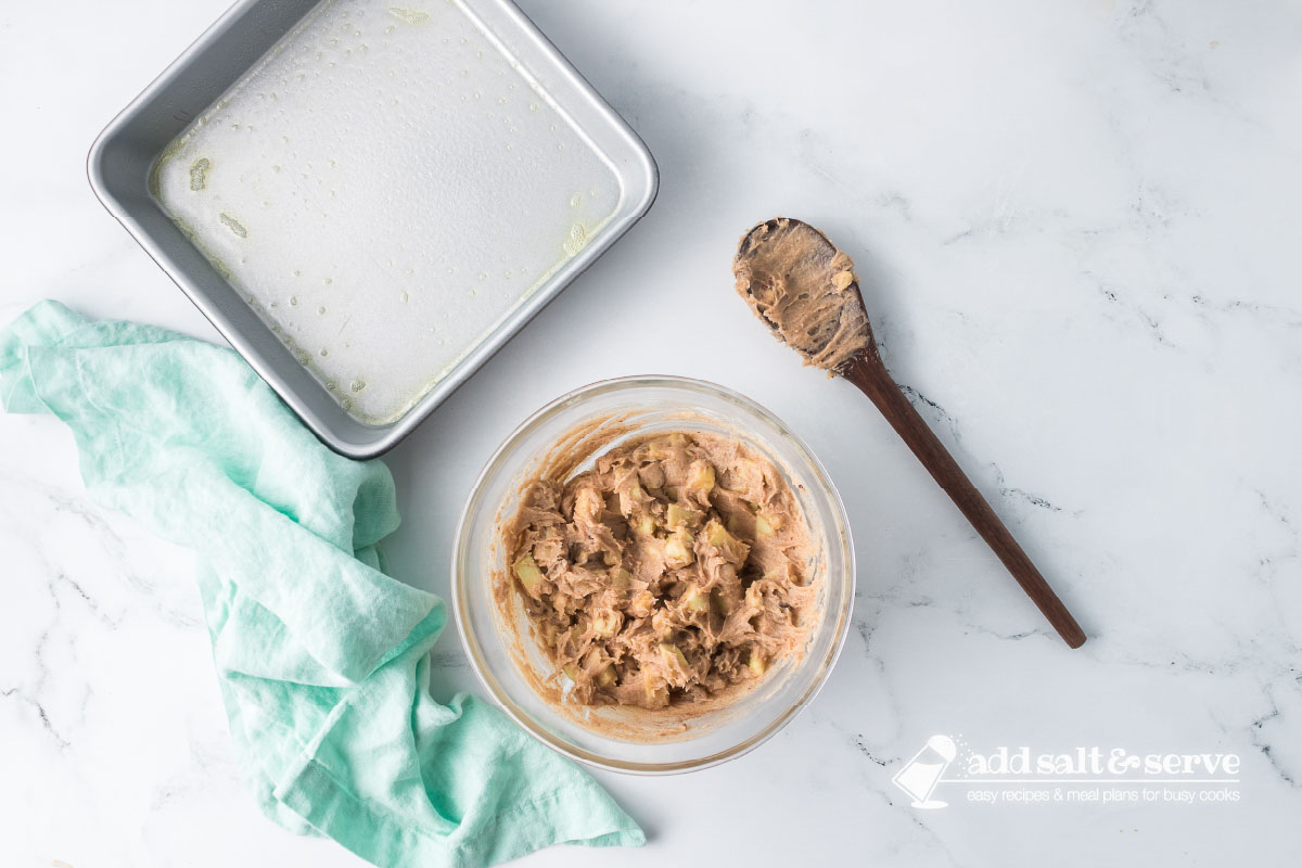 A clear glass bowl with mixed batter for Apple Walnut Bars and a 9-inch by 9-inch baking pan sprayed with non-stick cooking spray beside the bowl.