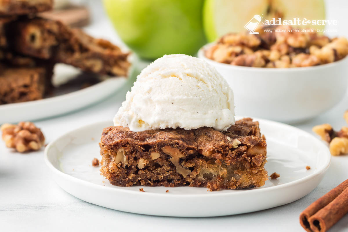 An Apple Walnut Bar topped with vanilla ice cream on a white plate with a green apple and a bowl of walnuts in the background.
