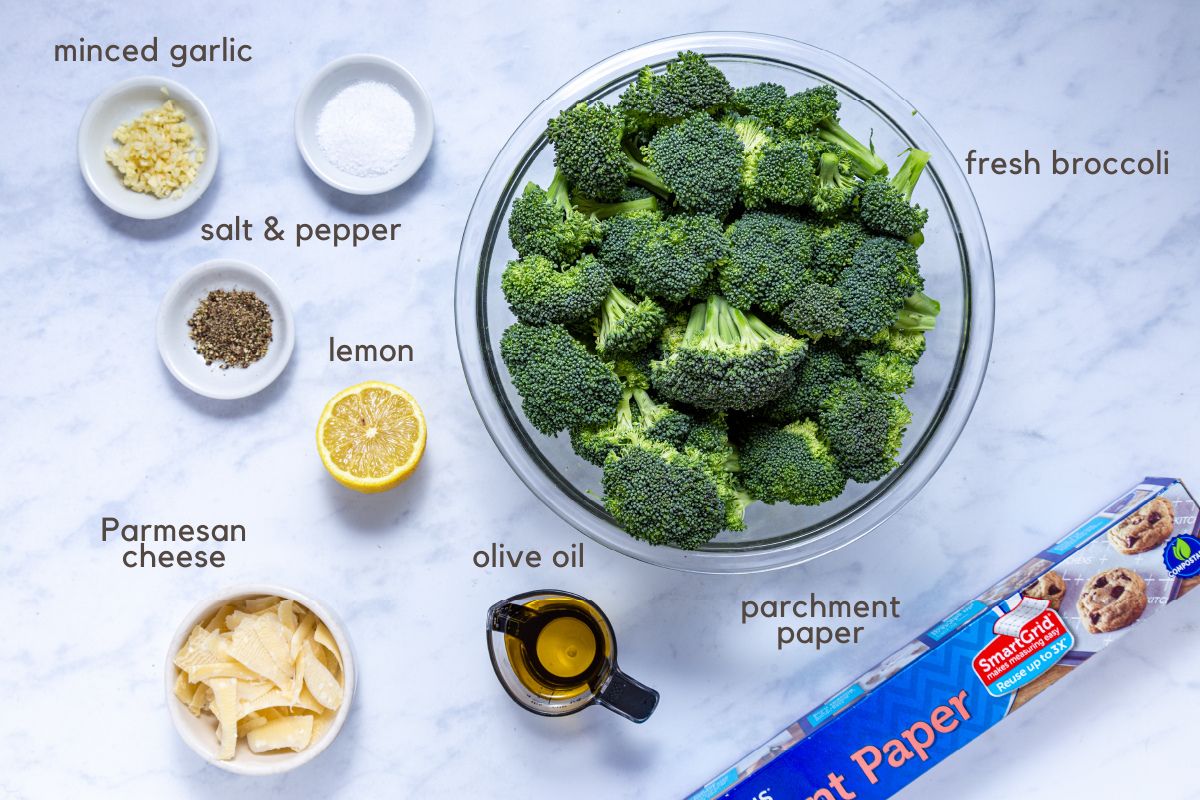 Ingredients for Roasted Garlic Broccoli: a large bowl of broccoli florets, small bowls of salt, pepper, garlic, and parmesan cheese, half a lemon, and a measuring cup with olive oil. A roll of parchment paper is beside the bowls.