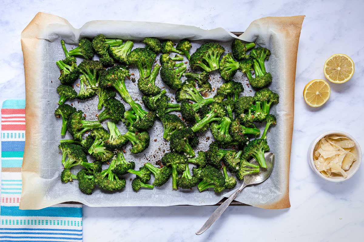 Roasted broccoli florets on a sheet pan lined with parchment paper. There is a spoon with the broccoli and a small bowl of parmesan cheese beside the pan. Two lemon halves are next to the bowl.