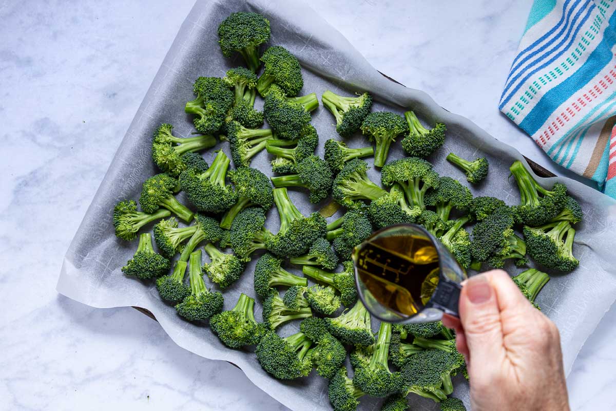 A person is using a small measuring cup to pour olive oil onto broccoli florets on a parchment-lined baking sheet