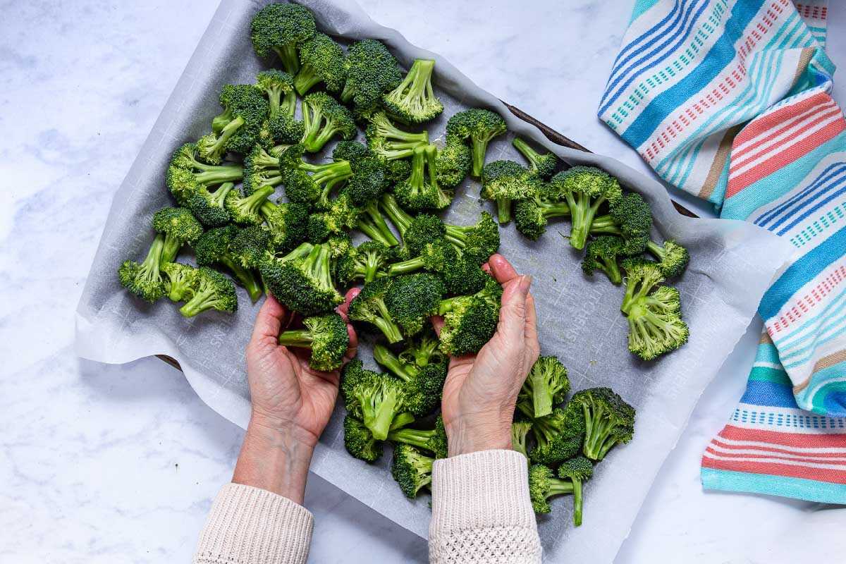 A person is using their hands to put broccoli florets on a baking sheet lined with parchment paper.