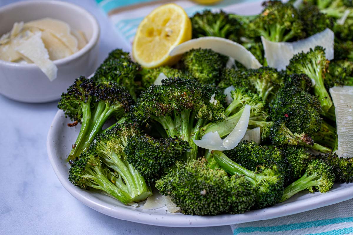 Broccoli florets on a plate with a lemon half. There's a small bowl of parmesan cheese beside the plate.