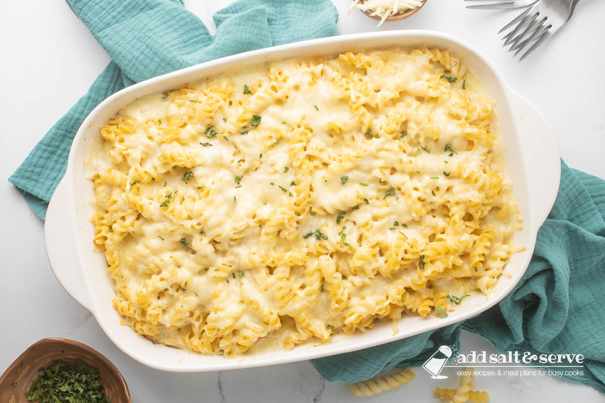 Rotini Pasta Bake in a baking dish resting on a tea towel with forks, shredded cheese, and chopped fresh parsley surrounding the pan.
