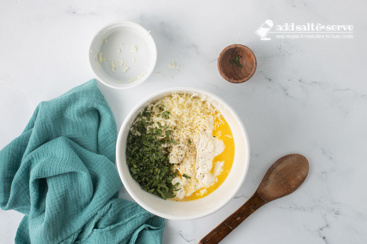 A medium-sized bowl with ricotta cheese, eggs, shredded mozzarella cheese, minced parsley, and salt and pepper before it has been mixed.