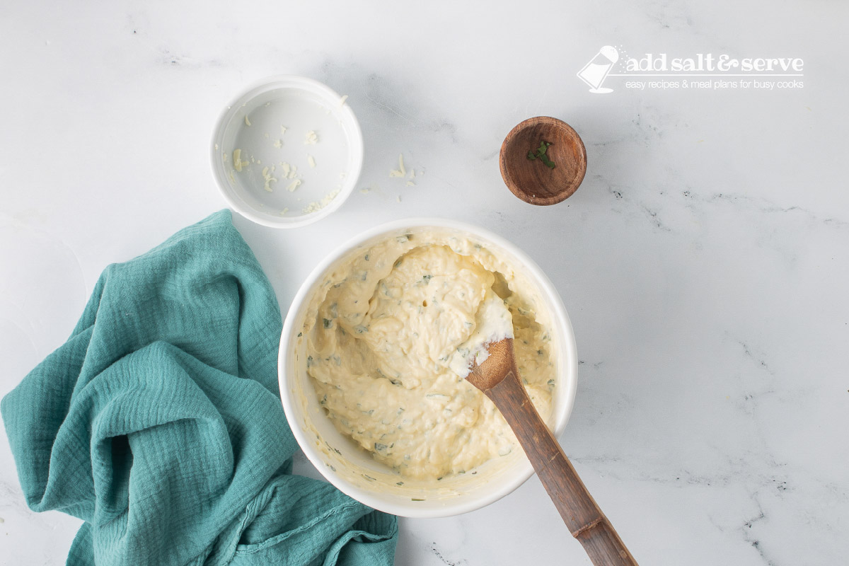 A medium-sized bowl with ricotta cheese, eggs, shredded mozzarella cheese, minced parsley, and salt and pepper after it has been mixed.