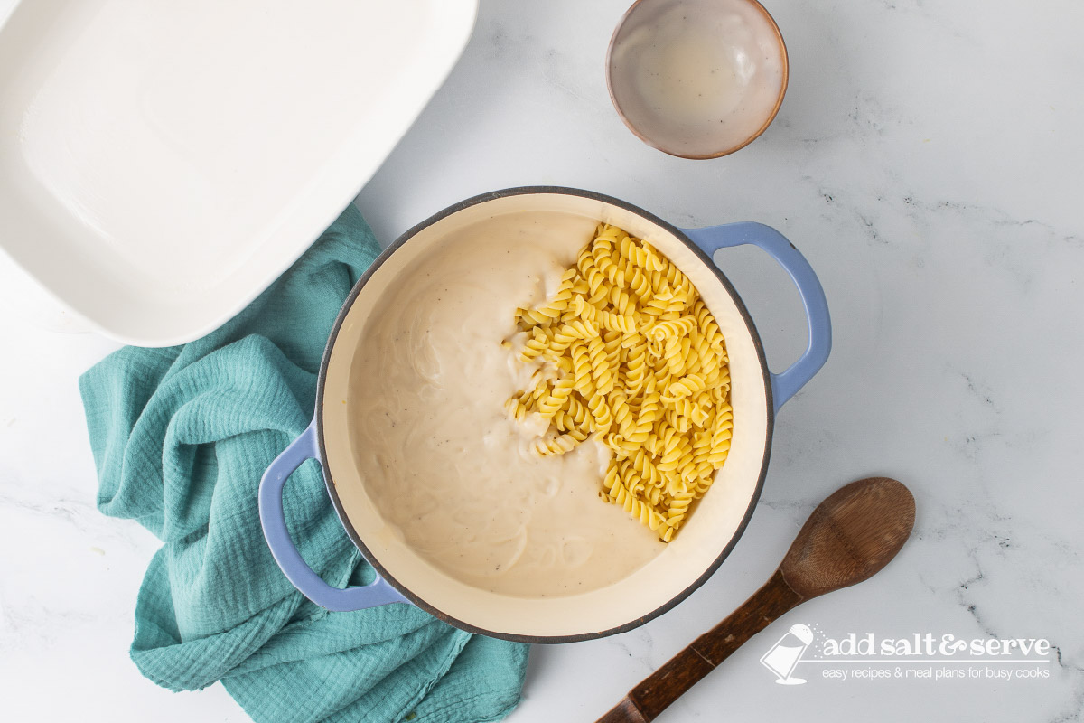 A Dutch oven with Alfredo sauce and cooked rotini pasta before it has been stirred.