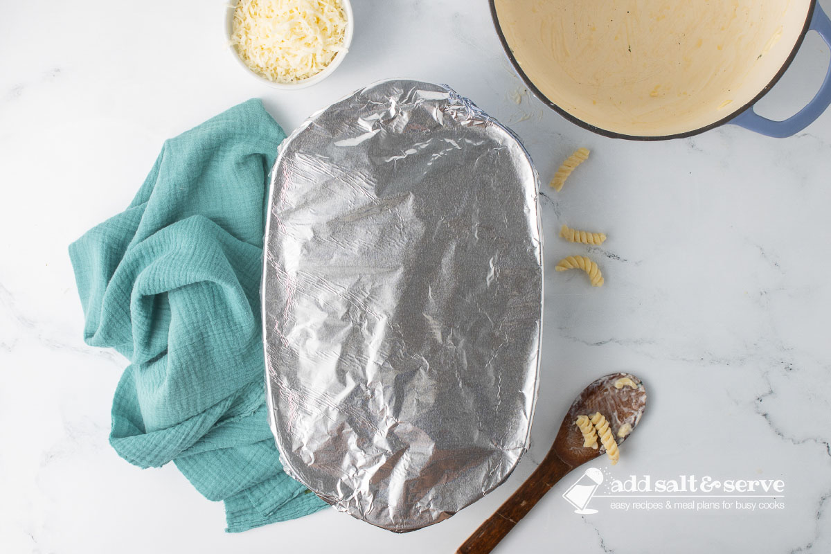 An oblong baking dish covered in aluminum foil on a counter beside an empty Dutch oven and a wooden spoon with saucy rotini on it.