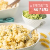 A bowl of Alfredo Rotini Pasta Bake with a fork surrounded by parsley, uncooked rotini, and a bowl of broccoli on a counter.